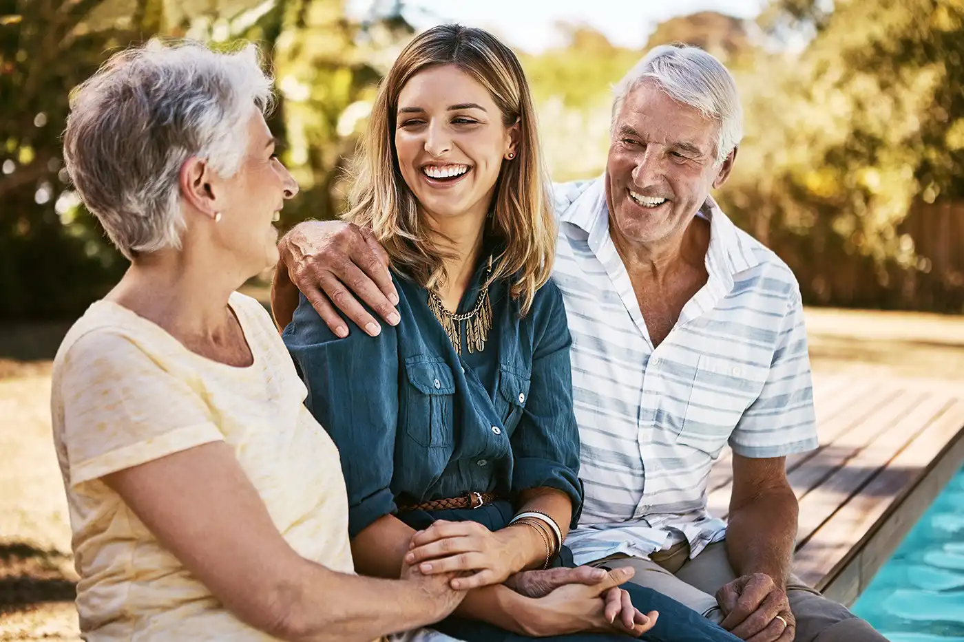 An adult daughter sits in-between her senior parents by the pool outside